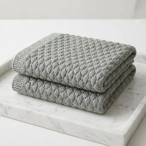 Puede incluir: Dos mantas grises tejidas y dobladas con un patr&oacute;n de diamantes apiladas sobre una bandeja de m&aacute;rmol blanco. Las mantas tienen una superficie texturizada y una apariencia suave, ideales para la decoraci&oacute;n del hogar.