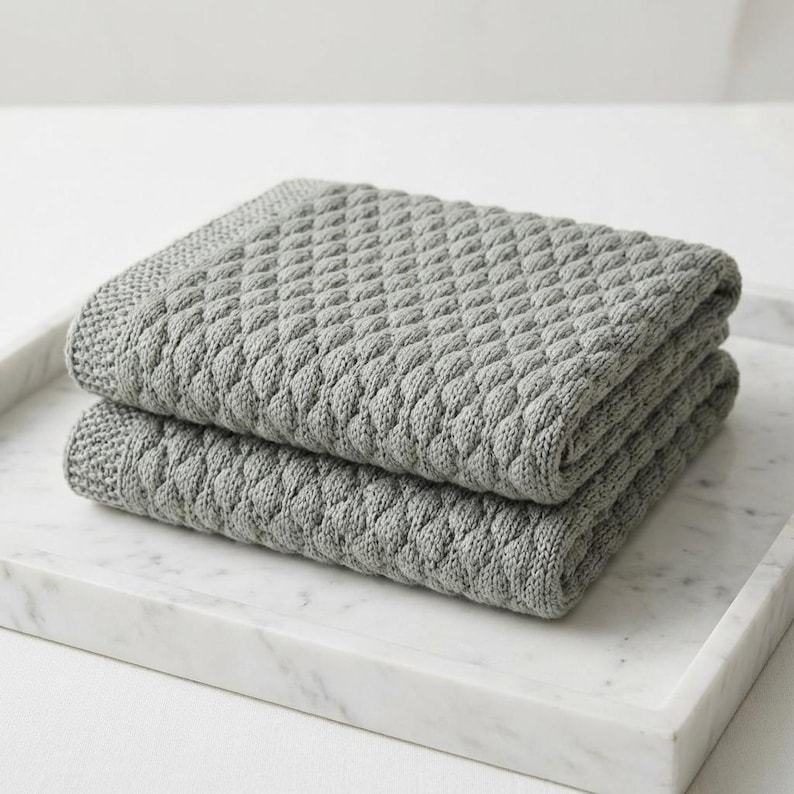 Puede incluir: Dos mantas grises tejidas y dobladas con un patr&oacute;n de diamantes apiladas sobre una bandeja de m&aacute;rmol blanco. Las mantas tienen una superficie texturizada y una apariencia suave, ideales para la decoraci&oacute;n del hogar.