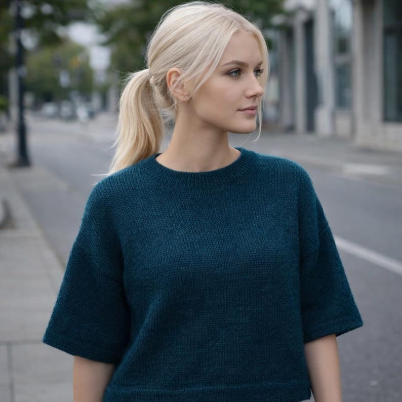 May include: A teal-colored sweater with short sleeves. The sweater has a crew neck and a slightly loose fit. The material appears to be a soft knit. The background shows a street scene with buildings and trees.