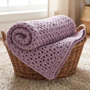 Peut inclure: Une couverture en crochet de couleur lavande, roulée, est posée dans un panier en osier brun. La couverture présente un motif ajouré. Le panier est posé sur un tapis clair et texturé, avec une fenêtre en arrière-plan.