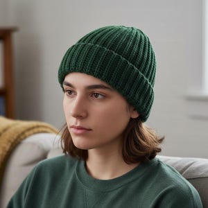 Op de afbeelding: Een donkergroene gebreide beanie. De hoed is gemaakt van een geribbelde gebreide stof en heeft een omgeslagen rand. De persoon op de foto draagt een groene trui. De hoed is een winteraccessoire.