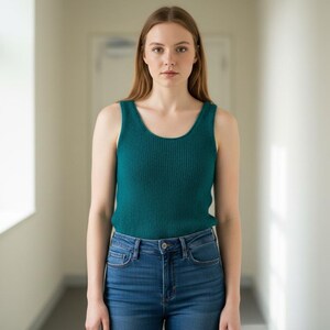 May include: A teal ribbed tank top and blue denim jeans. The tank top has a scoop neck and is made of a textured material. The jeans are a classic five-pocket style. The person is standing in a hallway with natural light.