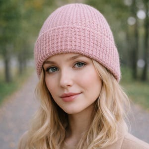 Puede incluir: Un gorro de punto acanalado de color rosa claro. El gorro está hecho de un material suave y lo lleva una persona con cabello rubio y ojos azules. El fondo es una escena borrosa al aire libre con árboles.