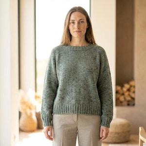 May include: A woman wearing a sage green knit sweater with a crew neck and long sleeves. The sweater has a speckled pattern and ribbed cuffs and hem. She is wearing beige pants. The background includes a window and a fireplace.