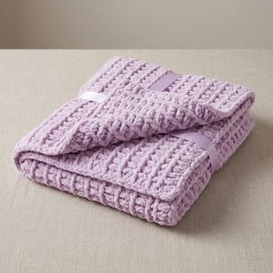 May include: A folded lavender blanket made of thick, plush yarn with a textured, waffle-like stitch pattern. The blanket is secured with a smooth, matching lavender satin ribbon tied around the center, creating a neat and organized presentation.