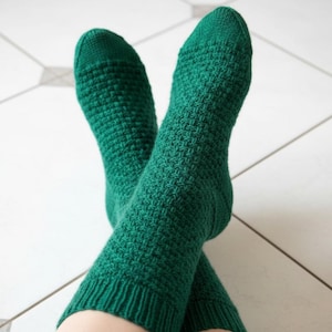 Könnte beinhalten: Ein Paar smaragdgrüne Strickstrümpfe mit strukturiertem Muster sind an den Knöcheln gekreuzt. Die Socken sind aus weichem Material und haben einen gerippten Bund. Der Hintergrund ist ein weißer Fliesenboden mit grauen Linien.