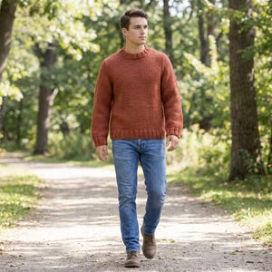 May include: A person wearing a rust-colored knit sweater and blue jeans walks on a path in a park. The sweater has a crew neck and ribbed cuffs. The jeans are a classic five-pocket style. The person is wearing brown leather shoes.