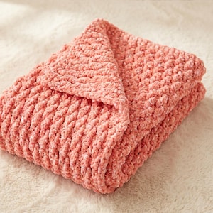 Puede incluir: Una manta plegada de color coral con un tejido de punto grueso y texturizado. La manta está hecha de un material suave y afelpado y está doblada cuidadosamente, mostrando su aspecto acogedor. La manta es de color liso y parece nueva.