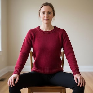 May include: A person wearing a burgundy crew neck sweater and black leggings sits on a wooden chair. The sweater has ribbed cuffs and a ribbed hem. The person is in a room with a window and a couch.