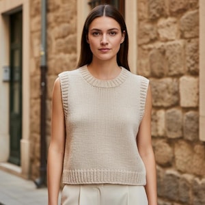 May include: A beige knitted sleeveless sweater with a ribbed crew neck and hem. The sweater is made of a soft, textured material. The model is wearing the sweater with beige pants. The background is a stone building.