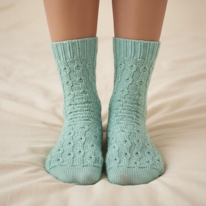 May include: A pair of light blue knitted socks with a textured pattern. The socks are worn on a person's feet, and the image is taken from a front-facing perspective. The socks are made of a soft material and are likely for warmth and comfort.