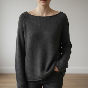 以下が含まれることがあります： ボートネックと長袖のダークグレーのセーター。セーターはリブ編みのテクスチャで、ゆったりとしたフィット感です。ダークグレーのパンツを着用しています。背景はプレーンな白い壁です。