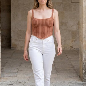 May include: A crocheted, rust-colored top with a square neckline and wide straps. The top is paired with white jeans. The model is walking in a stone-walled corridor. The top is a crop top style.