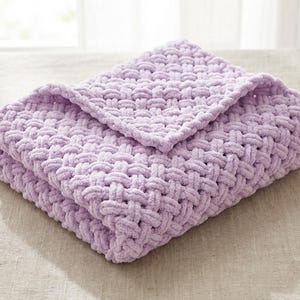 May include: A folded, lavender-colored blanket with a woven texture. The blanket is made of a soft, plush material and is folded neatly, with the top layer partially covering the bottom layer. The blanket is resting on a neutral-colored surface.
