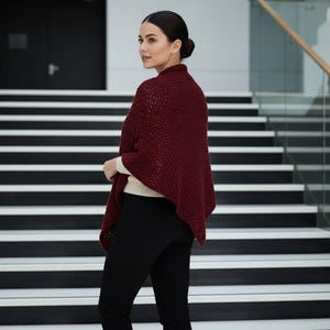 Może przedstawiać: Kobieta w bordowym dzianinowym szalu na kremowym topie i czarnych spodniach. Szal ma wzorzystą fakturę. Tło przedstawia schody z czarno-białymi stopniami i szklaną balustradą.