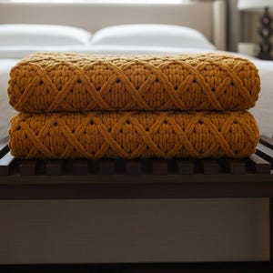 May include: Two mustard-yellow, chunky knit blankets are stacked on a dark brown wooden bench. The blankets have a diamond pattern and are placed in front of a bed with white linens. The room has a neutral colour palette.