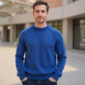 May include: A blue crew neck sweater. The sweater is a solid color and has long sleeves. The person is wearing the sweater and dark blue jeans. The background is a building with glass windows.