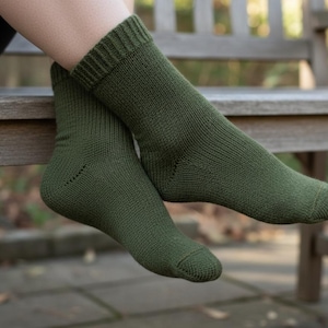 Op de afbeelding: Een paar olijfgroene sokken. De sokken zijn gebreid en hebben een geribbelde boord. De sokken worden gedragen op de voeten van een persoon, met een houten bank en groen in de achtergrond.