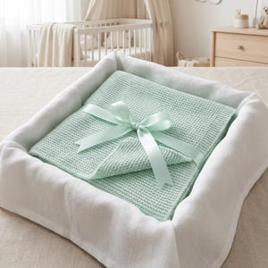 May include: A mint green knitted baby blanket, folded and tied with a matching satin ribbon bow, rests inside a white linen-lined basket. The blanket is a soft, textured knit, perfect for a nursery setting.