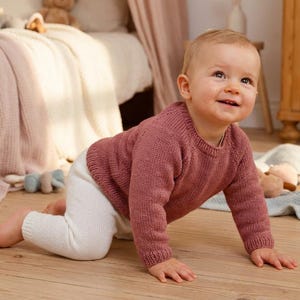 May include: A baby wearing a rose-colored knit sweater and white pants crawls on a wooden floor. The baby is smiling. A bed with a pink blanket and stuffed animals are in the background.