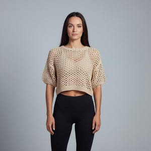 May include: A beige, open-weave crop top with short sleeves. The top is paired with black leggings. The model is standing in front of a grey background. The top's texture is visible.