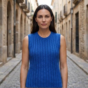 May include: A sleeveless, ribbed knit top in a vibrant blue hue. The top has a crew neck and a close fit, showcasing a vertical ribbing pattern. The model is standing in a cobblestone street.