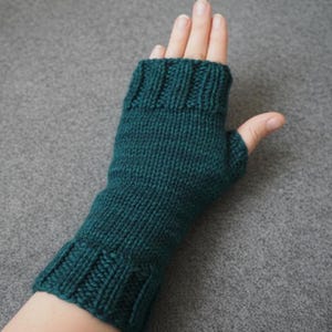 May include: A teal green knitted fingerless glove. The glove is made of a textured yarn and features a ribbed cuff and a thumb opening. The glove is designed to cover the hand and wrist, leaving the fingers free.