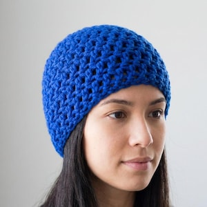 Op de afbeelding: Een close-up van een felblauwe gehaakte beanie. De hoed is gemaakt met een getextureerde steek, waardoor een ademende, open weefsel ontstaat. De beanie is effen van kleur en lijkt handgemaakt. De achtergrond is een zachte, neutrale tint.