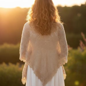Puede incluir: Una persona con un chal beige con un diseño geométrico y bordes festoneados, de pie al aire libre. El chal está sobre un vestido blanco. El fondo muestra un paisaje verde borroso y una cálida puesta de sol.