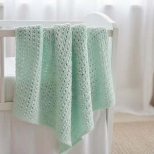 May include: A pale green crocheted baby blanket draped over a white cot. The blanket features a textured, open weave pattern. The cot has a white skirt and is set against a soft, neutral background.