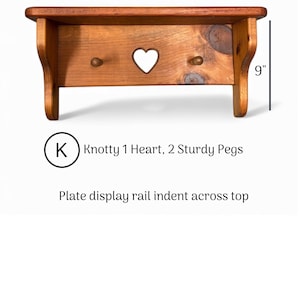 May include: A brown wooden wall shelf with a plate display rail indent across the top, featuring a central heart cutout and two sturdy pegs. The shelf measures 18 inches wide by 9 inches high, labeled as Knotty 1 Heart, 2 Sturdy Pegs.