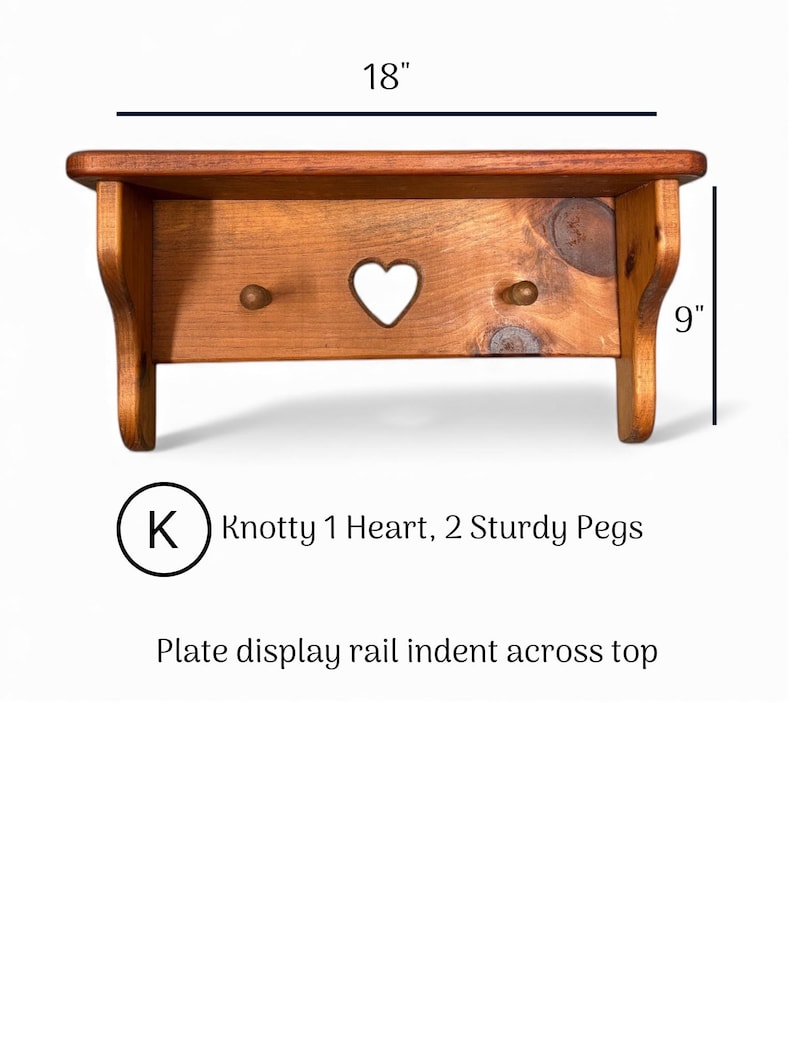 May include: A brown wooden wall shelf with a plate display rail indent across the top, featuring a central heart cutout and two sturdy pegs. The shelf measures 18 inches wide by 9 inches high, labeled as Knotty 1 Heart, 2 Sturdy Pegs.