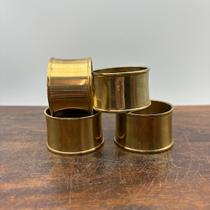May include: Four gold-colored napkin rings stacked on a wooden surface. The rings are cylindrical with a smooth, polished finish. The warm tones of the gold contrast with the wood grain.