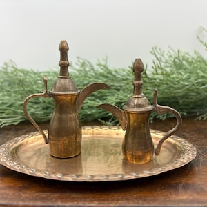 May include: Two antique brass coffee pots with curved spouts and handles sit on a decorative brass tray. The tray has a detailed edge. The background includes green foliage and a dark wooden surface.
