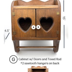 May include: A brown wooden wall cabinet with two doors featuring heart-shaped cutouts and white knobs. It includes a towel rod at the bottom and measures 12 inches wide, 15 inches high, and 4.5 inches deep. Text reads: G Cabinet w/ Doors and Towel Rod.