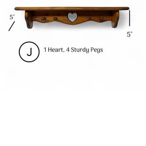 May include: A brown wooden wall shelf featuring a heart-shaped cutout in the center and four sturdy pegs. The shelf measures 24 inches in width and 5 inches in height. Text on the image reads: J 1 Heart, 4 Sturdy Pegs.