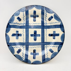 May include: A round ceramic plate with a blue and white geometric pattern. The design features blue crosses, wavy lines, and squares. The plate has a dark blue rim and a cream-colored background.