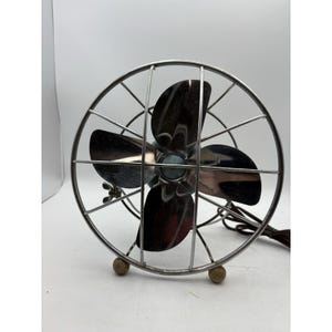 May include: A vintage chrome-plated electric fan with four blades, enclosed by a wire cage. The fan has a round base with two small wooden feet. The power cord is brown. The fan is designed for tabletop use.