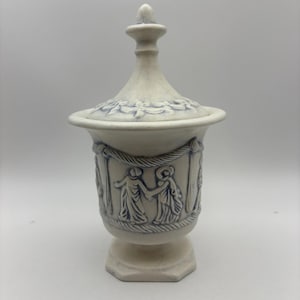 May include: A white ceramic urn with a lid, featuring blue relief designs. The urn has a hexagonal base and a detailed frieze of figures holding hands. The lid is topped with a small finial, and the overall design is classical.