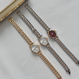 Puede incluir: Tres relojes de mujer vintage con esferas ovaladas y correas de metal. Un reloj tiene una esfera burdeos, mientras que los otros tienen esferas blancas. Las correas son doradas y plateadas, y los relojes están dispuestos en diagonal.