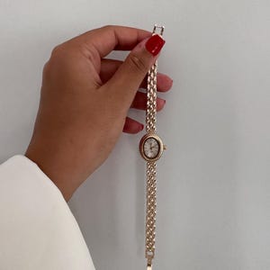 Op de afbeelding: Een goudkleurig polshorloge met een ovale wijzerplaat en een schakelband. De wijzerplaat van het horloge is wit met zwarte cijfers en wijzers. De band is gemaakt van kleine, verbonden schakels. Het horloge wordt tegen een witte achtergrond gehouden.