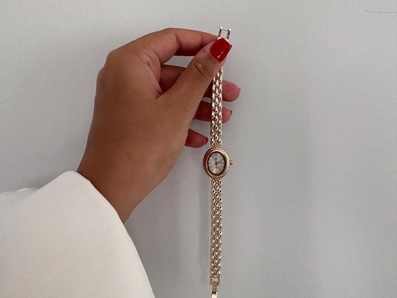 Op de afbeelding: Een goudkleurig polshorloge met een ovale wijzerplaat en een schakelband. De wijzerplaat van het horloge is wit met zwarte cijfers en wijzers. De band is gemaakt van kleine, verbonden schakels. Het horloge wordt tegen een witte achtergrond gehouden.