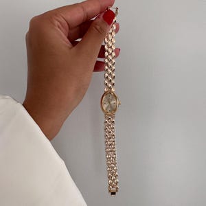 Op de afbeelding: Een goudkleurig polshorloge met een ovale wijzerplaat en een schakelband. Het horloge heeft een lichtgekleurde wijzerplaat met goudkleurige wijzers en markeringen. De band is gemaakt van kleine, afgeronde schakels. Het horloge wordt tegen een witte achtergrond gehouden.