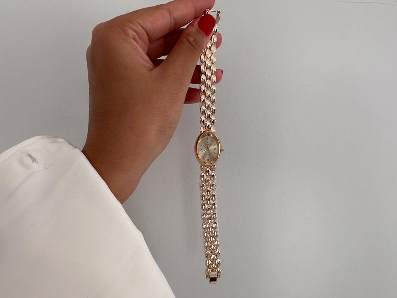 Op de afbeelding: Een goudkleurig polshorloge met een ovale wijzerplaat en een schakelband. Het horloge heeft een lichtgekleurde wijzerplaat met goudkleurige wijzers en markeringen. De band is gemaakt van kleine, afgeronde schakels. Het horloge wordt tegen een witte achtergrond gehouden.