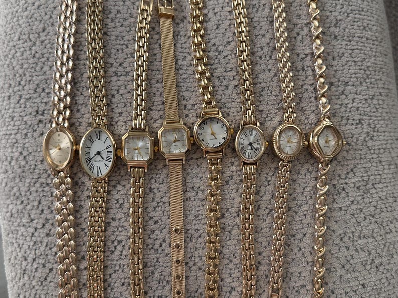 Op de afbeelding: Een verzameling goudkleurige polshorloges met verschillende wijzerplaatvormen en bandstijlen. De horloges hebben ovale, rechthoekige en ronde wijzerplaten met Romeinse cijfers. De bandjes omvatten schakel- en mesh-ontwerpen.