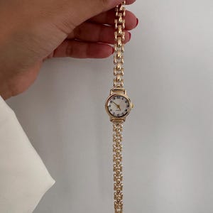 Op de afbeelding: Een goudkleurig polshorloge met een ronde wijzerplaat en een schakelband. De wijzerplaat heeft een witte achtergrond met zwarte cijfers en wijzers. De band is gemaakt van kleine, rechthoekige schakels.