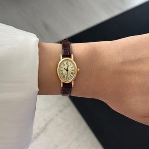 May include: A gold-toned wristwatch with an oval face and Roman numeral hour markers. The watch has a brown leather band. The watch face is approximately 0.75 inches wide.