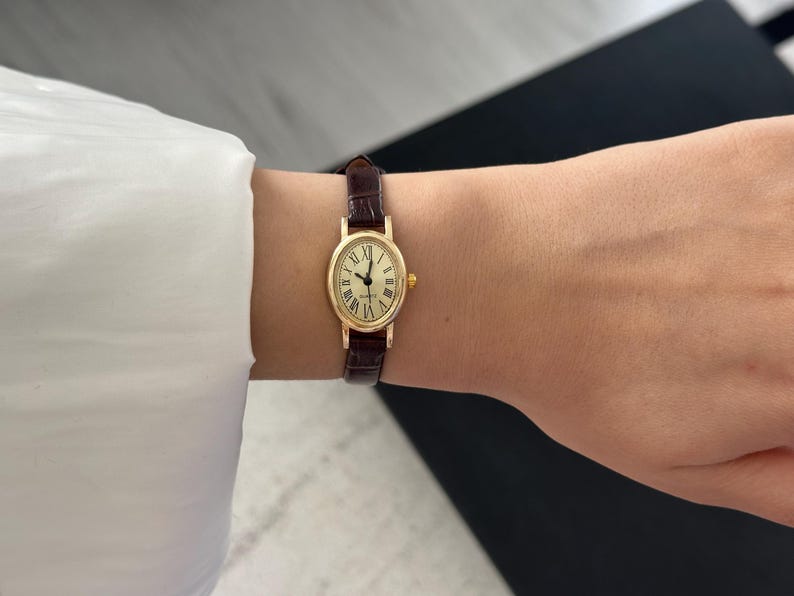 May include: A gold-toned wristwatch with an oval face and Roman numeral hour markers. The watch has a brown leather band. The watch face is approximately 0.75 inches wide.