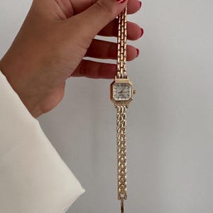 Op de afbeelding: Goudkleurig polshorloge met een rechthoekige wijzerplaat en een schakelband. Het horloge heeft een witte wijzerplaat met zwarte Romeinse cijfers en gouden wijzers. De band is gemaakt van kleine, rechthoekige schakels.