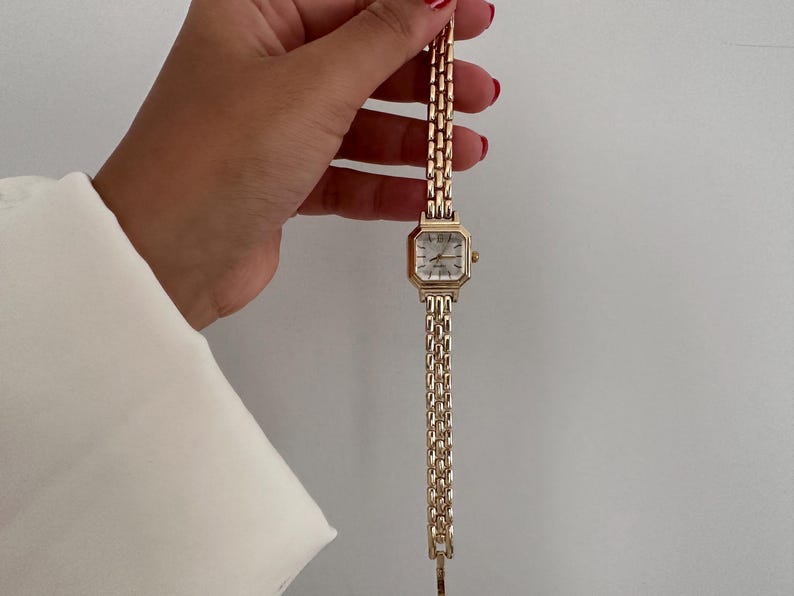 Op de afbeelding: Goudkleurig polshorloge met een rechthoekige wijzerplaat en een schakelband. Het horloge heeft een witte wijzerplaat met zwarte Romeinse cijfers en gouden wijzers. De band is gemaakt van kleine, rechthoekige schakels.